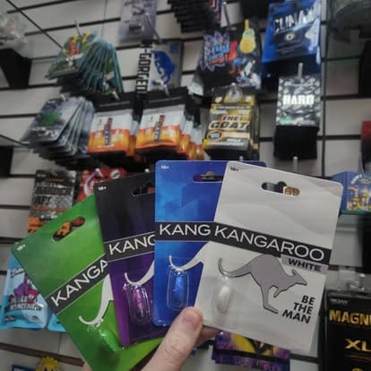 A hand holding four colorful single-pack retail cards of Kangaroo sexual wellness capsules against a store display background. The blister packs include Kangaroo Green, Violet, Blue, and White.