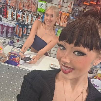 The store employee Liv (background) and her coworker Rem (foreground) smile for a casual selfie behind the checkout counter inside Paradise For Adults in Centralia, Washington, framed by walls lined with packaged novelty items.