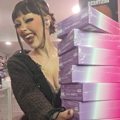 Rem Paradise smiling and sticking out her tongue while holding a tall stack of purple and pink holographic boxes inside the Paradise for Adults store in Centralia, Washington.